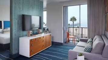 A hotel room with a large window, a flat-screen TV, a wooden dresser, a sofa, and a small table with a plant.