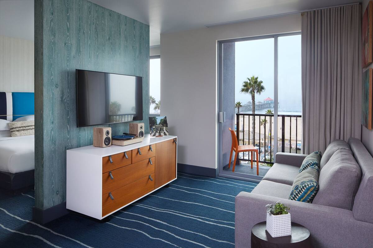 A modern hotel room with a large flat-screen TV, a wooden dresser, a grey sofa, and a balcony with a view of the beach.