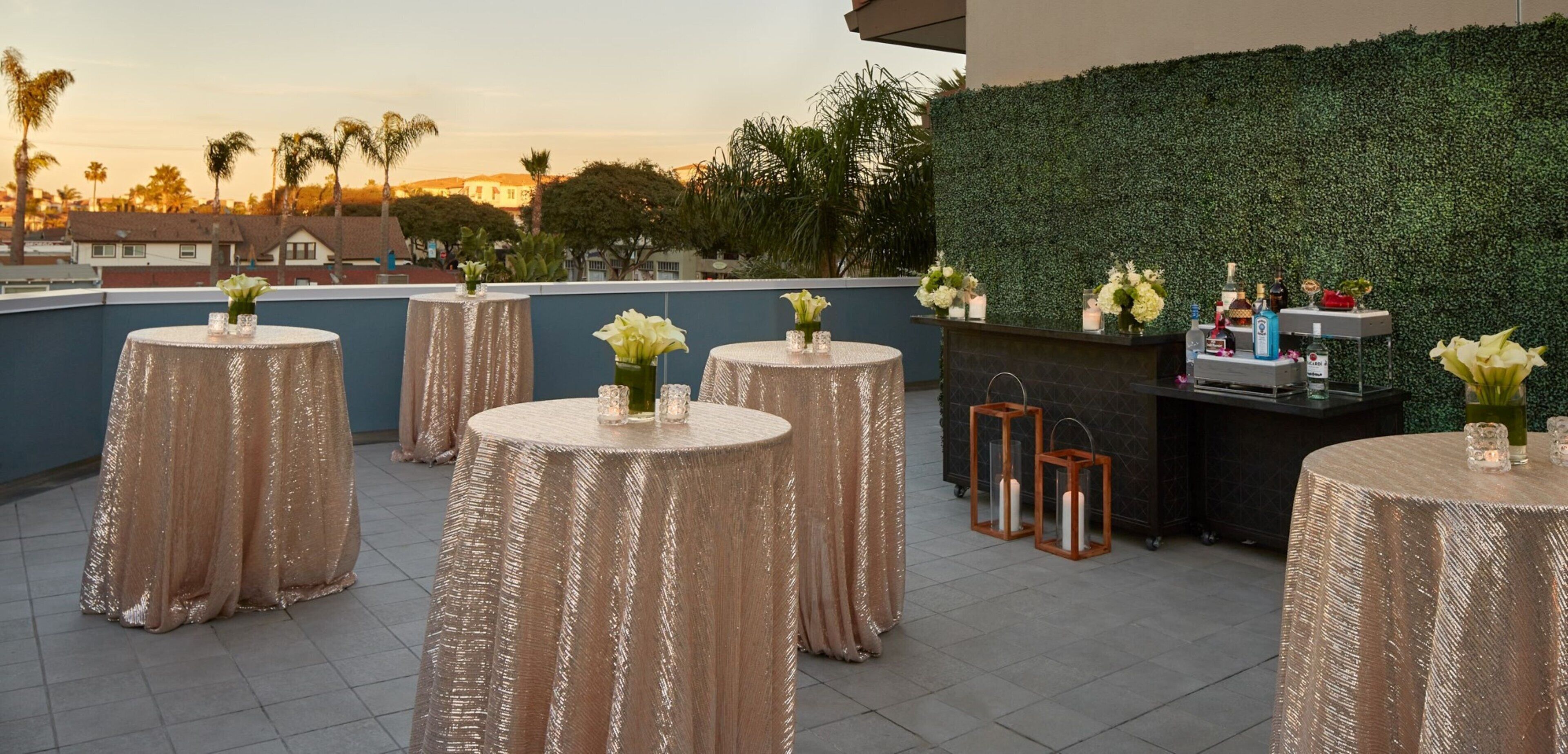 A rooftop terrace with round tables covered in glittery tablecloths, each with a vase of flowers and candles. A bar area with a green hedge backdrop and various bottles and glasses is visible.