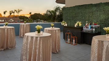 A rooftop terrace with round tables covered in glittery tablecloths, each with a vase of flowers and candles. A bar area with a green hedge backdrop and various bottles and glasses is visible.