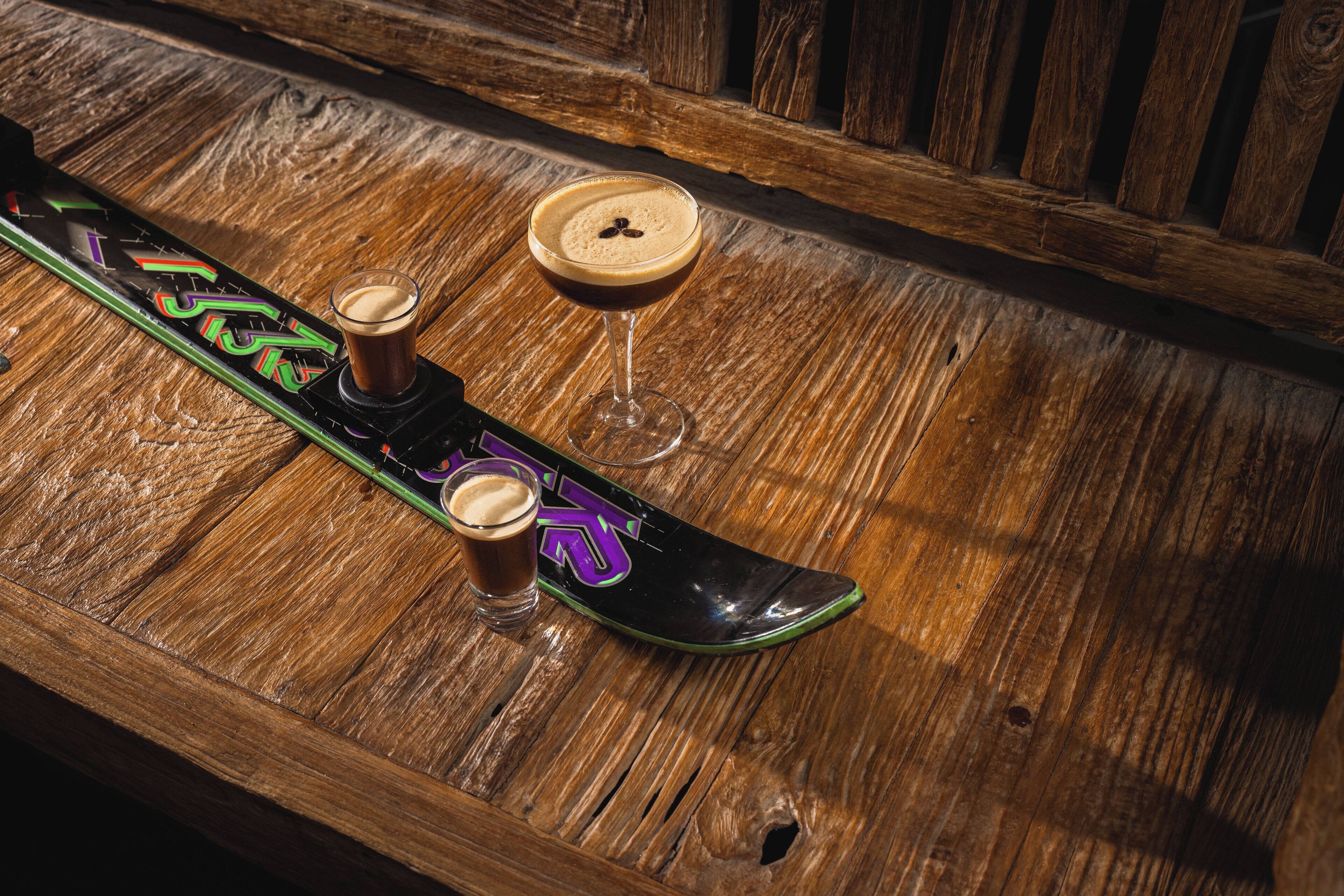 A wooden table with a snowboard and three glasses of a dark beverage.