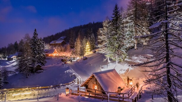 Una cabaña cubierta de nieve con fachada de madera, un pequeño porche y luces encendidas en el interior.
