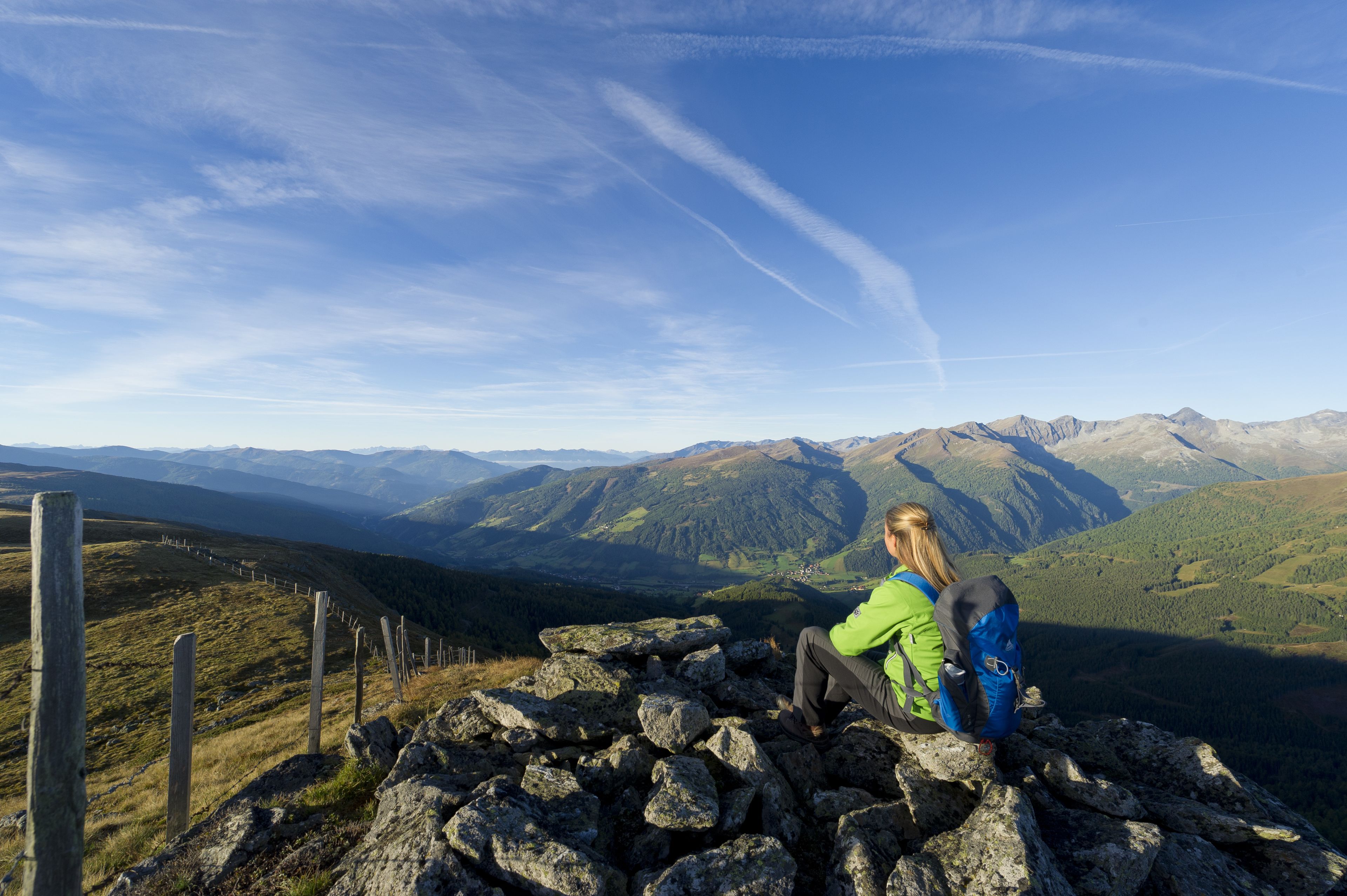 A person sitting on a rocky outcrop overlooking a mountainous landscape with a clear sky.
