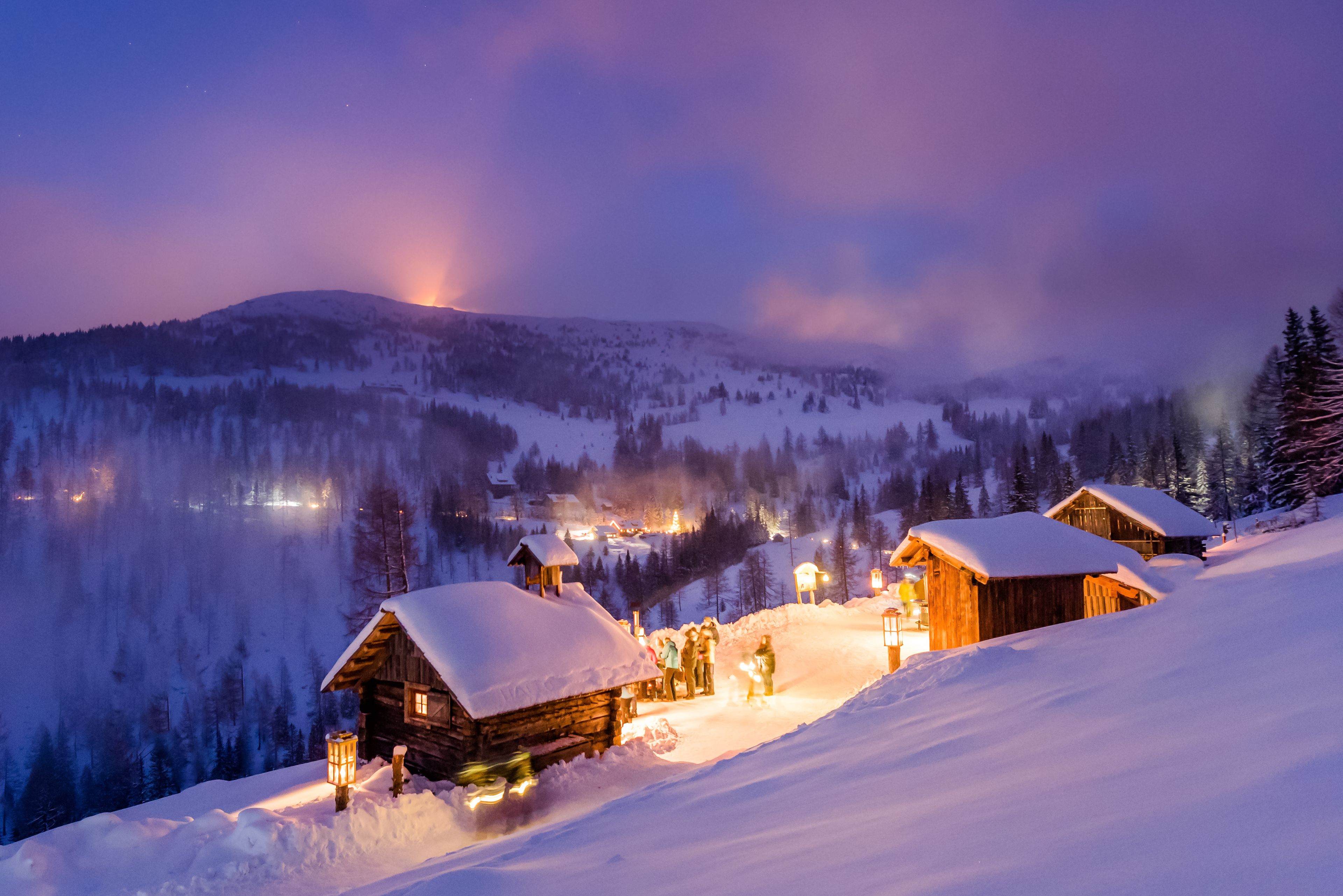A snow-covered mountain village at night with wooden cabins and a warm glow from the windows.