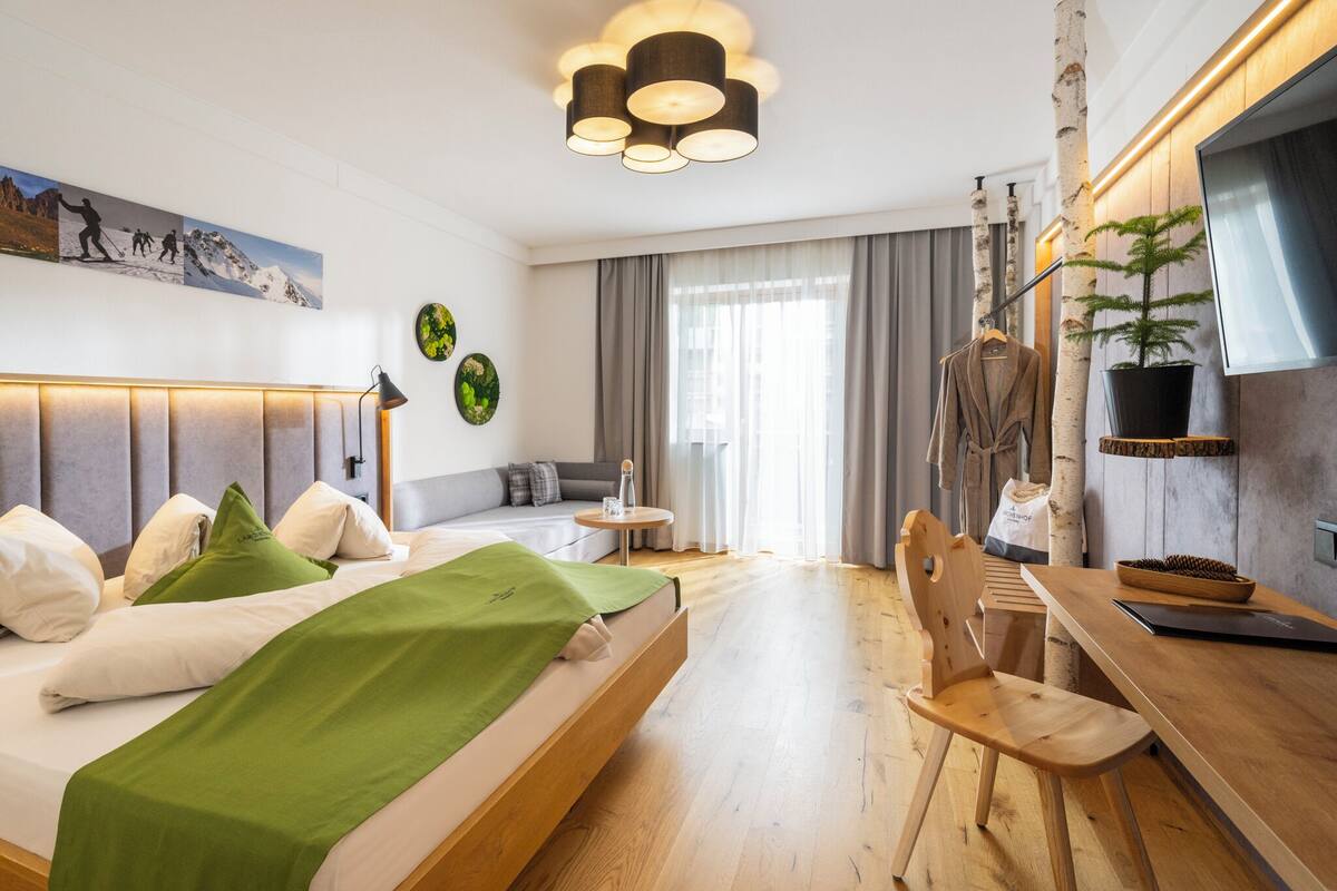 A modern hotel room with a bed, a desk, a chair, a plant, and a painting on the wall.