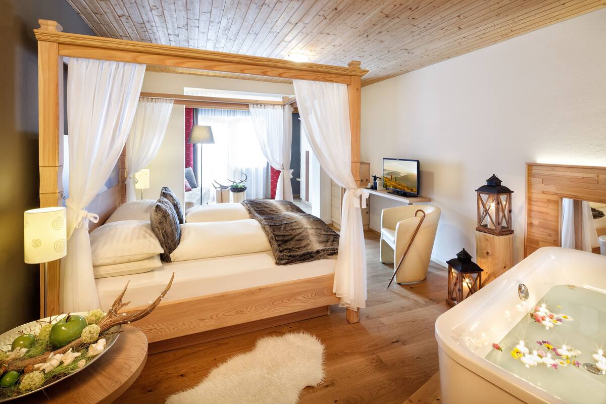 A bedroom with a four-poster bed, a sitting area with a chair, a small table, a decorative plant, and a bathtub with flowers.