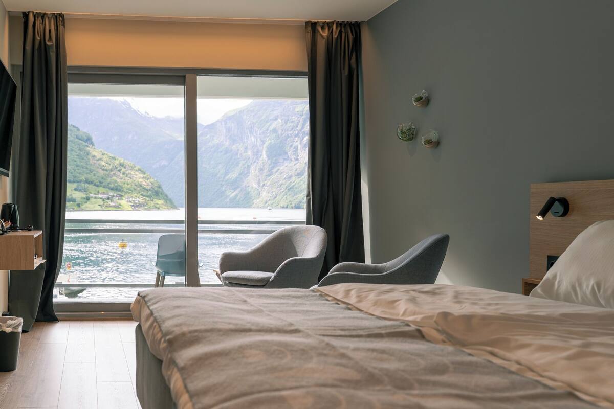 A hotel room with a bed, a chair, a small table, and a view of a lake and mountains.