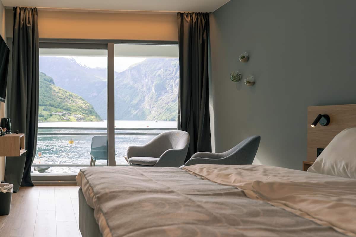 Habitación de hotel con cama, una silla, una mesita y vistas a un lago y montañas.