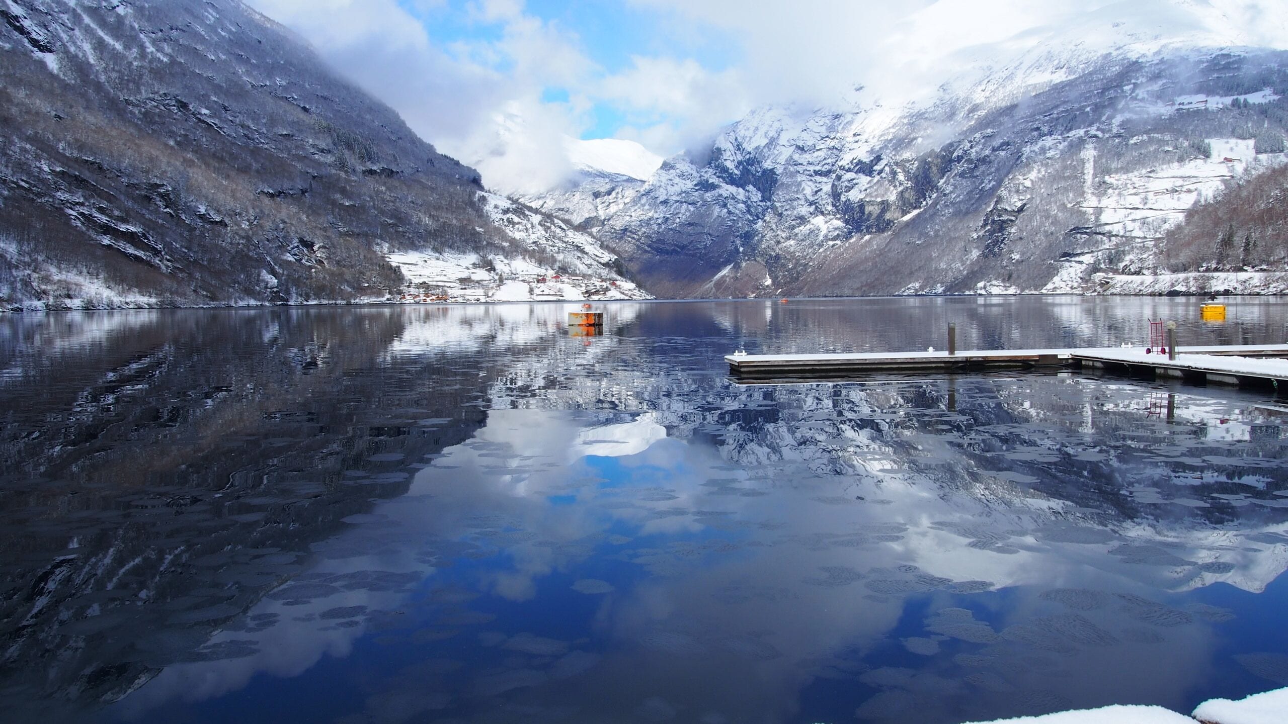 Un lago tranquilo rodeado de montañas nevadas, un muelle que se adentra en el agua y un cielo despejado.
