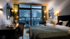 A hotel room with a bed, a desk with a chair, a small table, and a sofa. There are two windows with curtains, and a view of a snowy mountain. - Havila Hotel Geiranger (Stranda)
