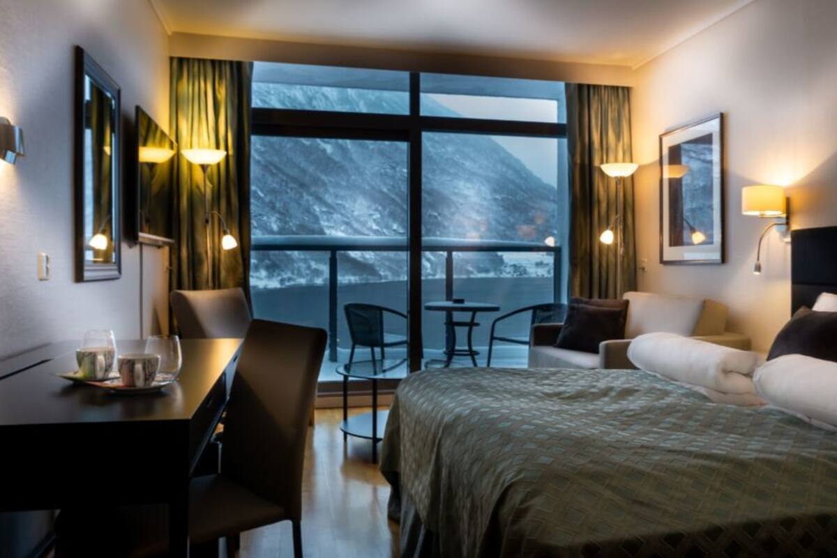 A hotel room with a bed, a desk with a chair, a small table, and a sofa. There are two windows with curtains, and a view of a snowy mountain.