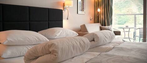 A hotel room with a large bed, multiple pillows, a headboard, bedside lamps, a framed picture, and a view of a mountainous landscape through a window.