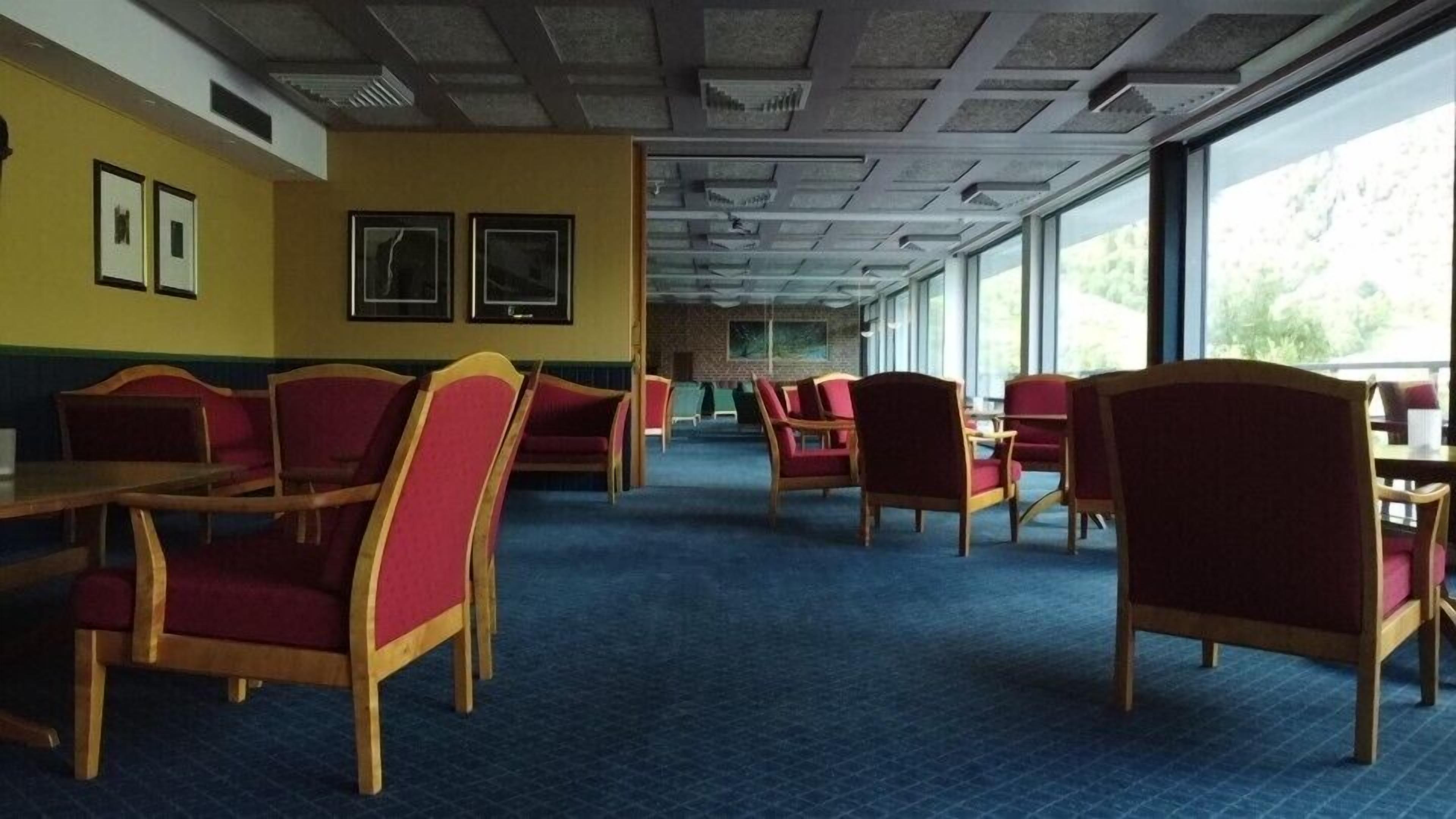 Un comedor con mesas de madera, sillas rojas, ventanas amplias y una alfombra azul.
