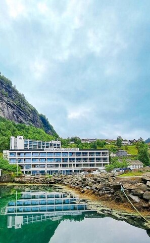 A modern hotel with multiple floors, a reflection in the water, and a mountainous backdrop. - Havila Hotel Geiranger (Stranda)