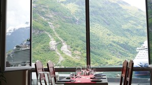 A dining table with chairs and wine glasses, set against a window with a view of a mountainous landscape and a cruise ship. - Havila Hotel Geiranger (Stranda)