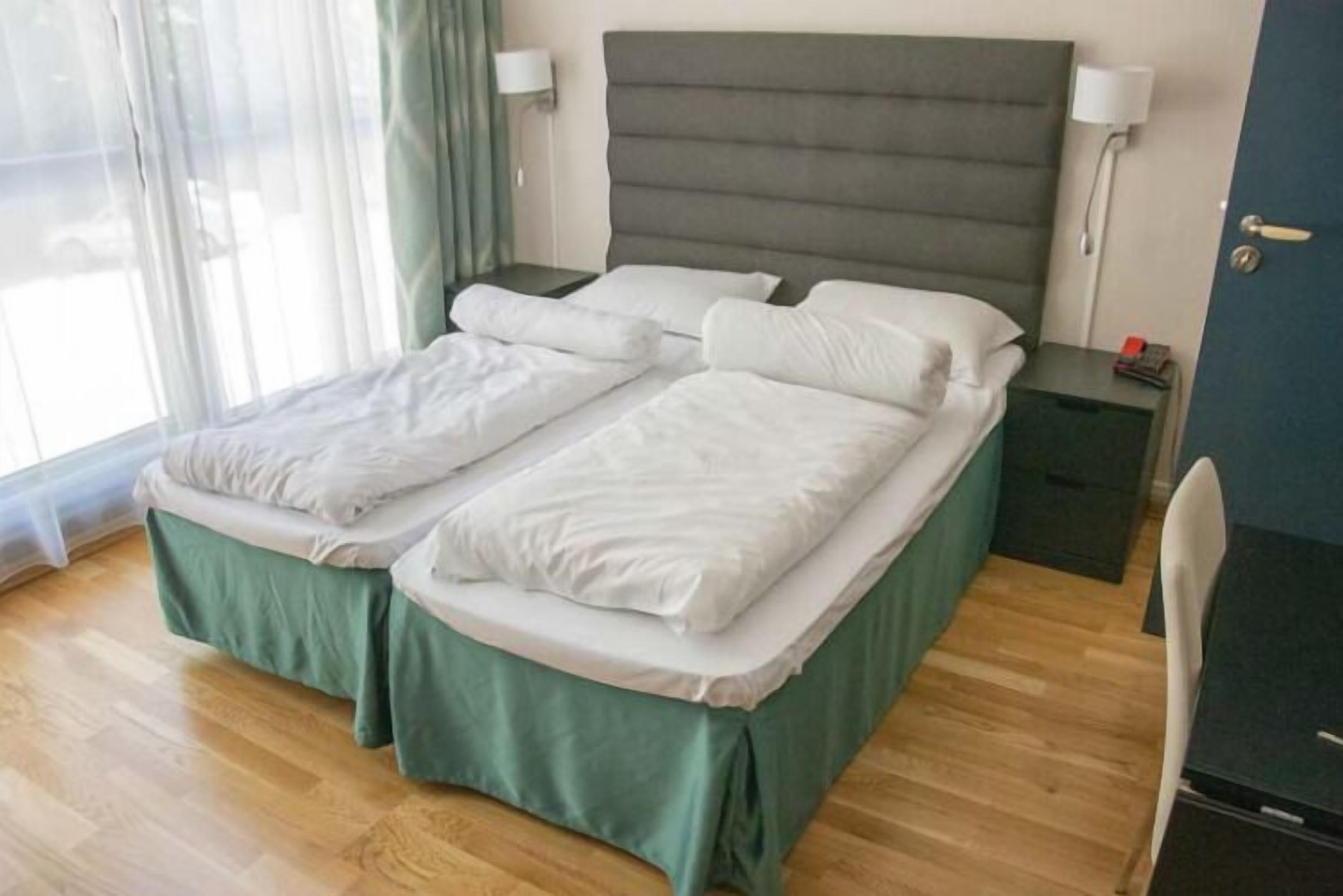 A double bed with a green bed skirt, two bedside tables with lamps, and a view of the street through a window with curtains.