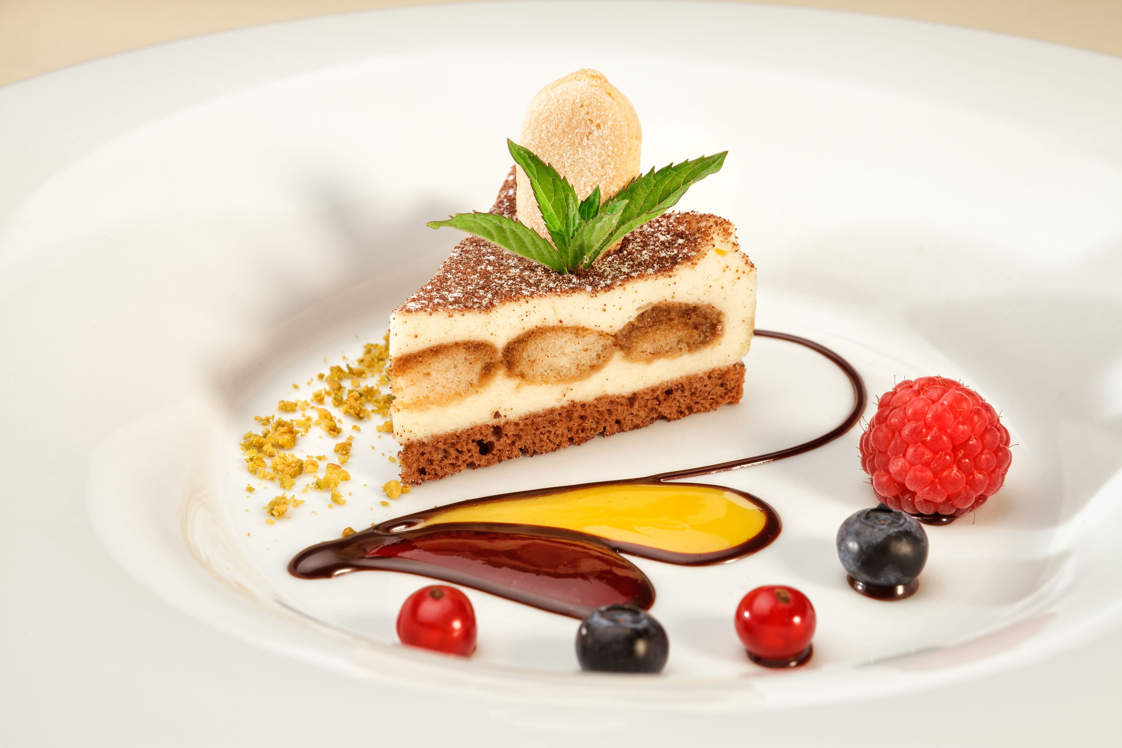 A slice of layered cake with a scoop of ice cream on top, garnished with mint leaves, and surrounded by berries and chocolate sauce.
