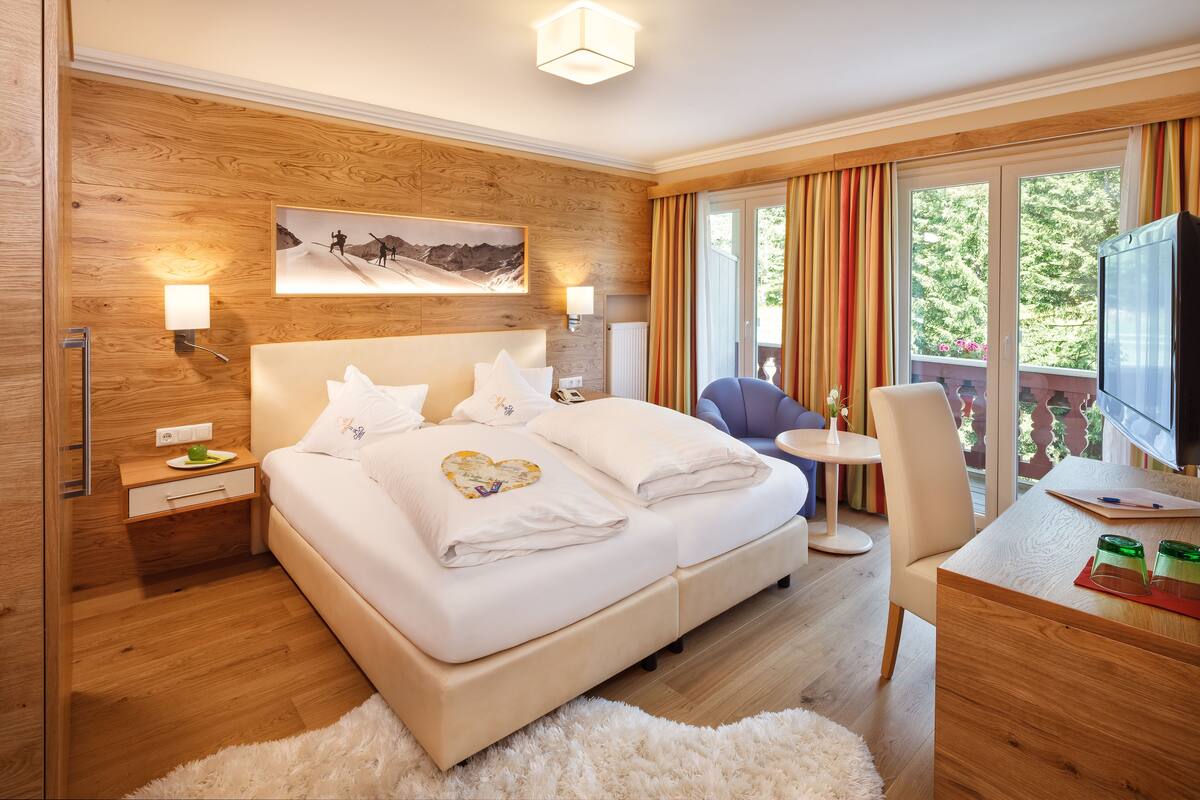 Habitación de hotel moderna con una pared de acento de madera, una cama grande con ropa blanca, un escritorio con televisor y un balcón con cortinas.