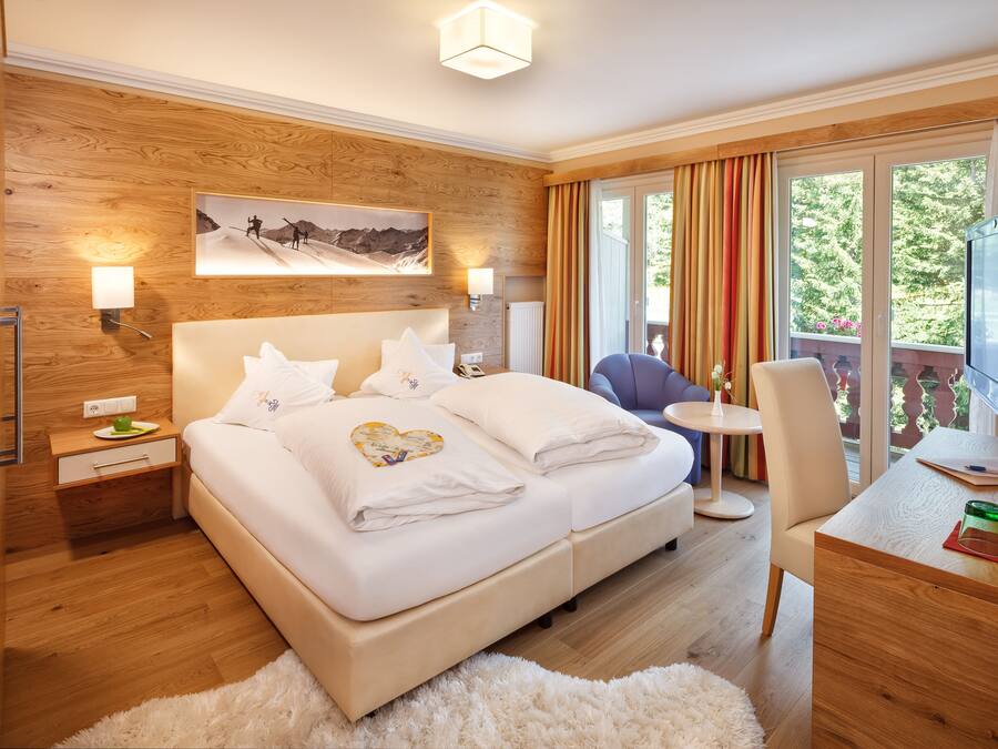 A modern hotel room with a wooden accent wall, a large bed with white linens, a desk with a television, and a balcony with curtains.