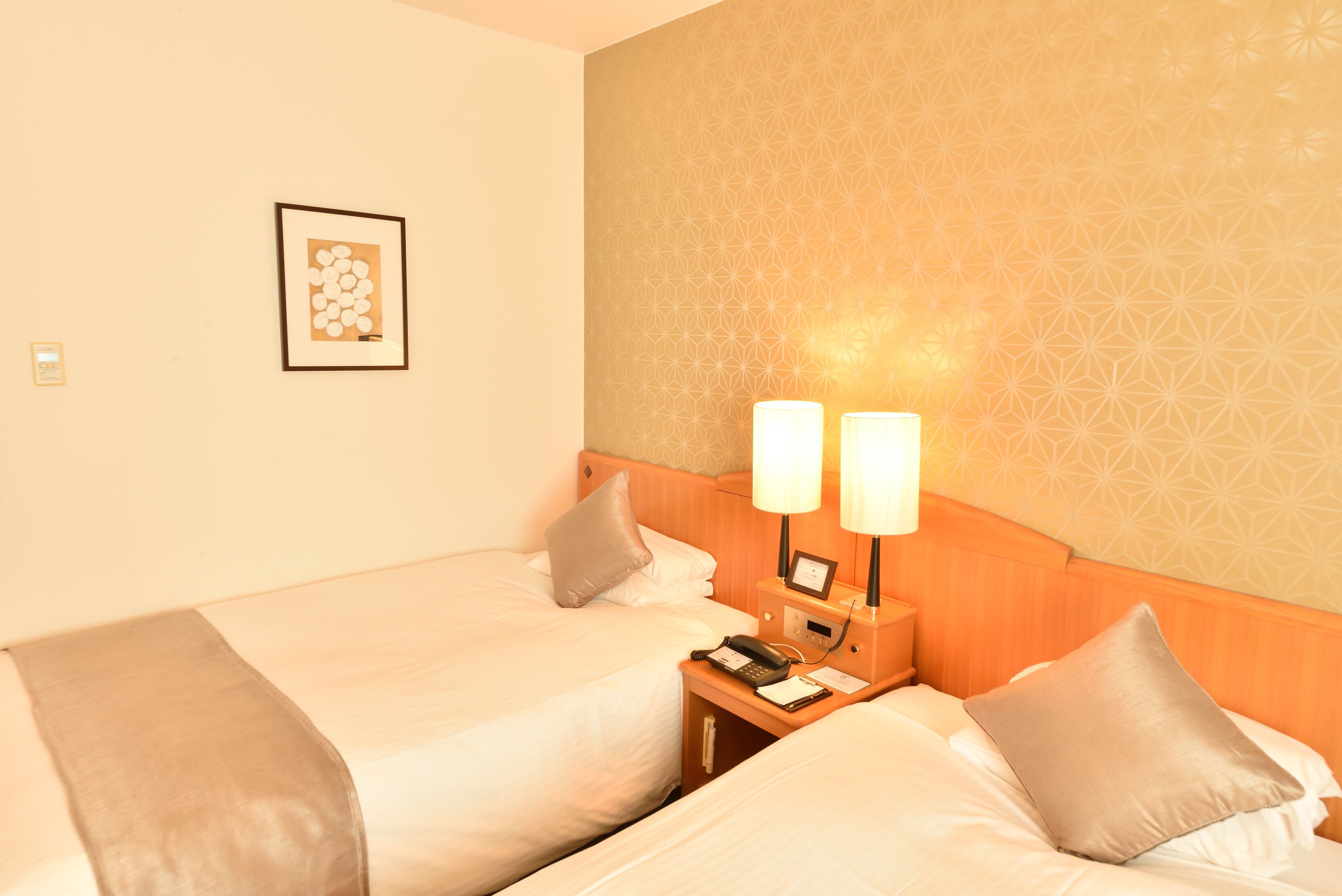 Ein Hotelzimmer mit zwei Betten, einem Nachttisch mit Lampe, einem gerahmten Bild an der Wand und einem gemusterten Wandpapier.