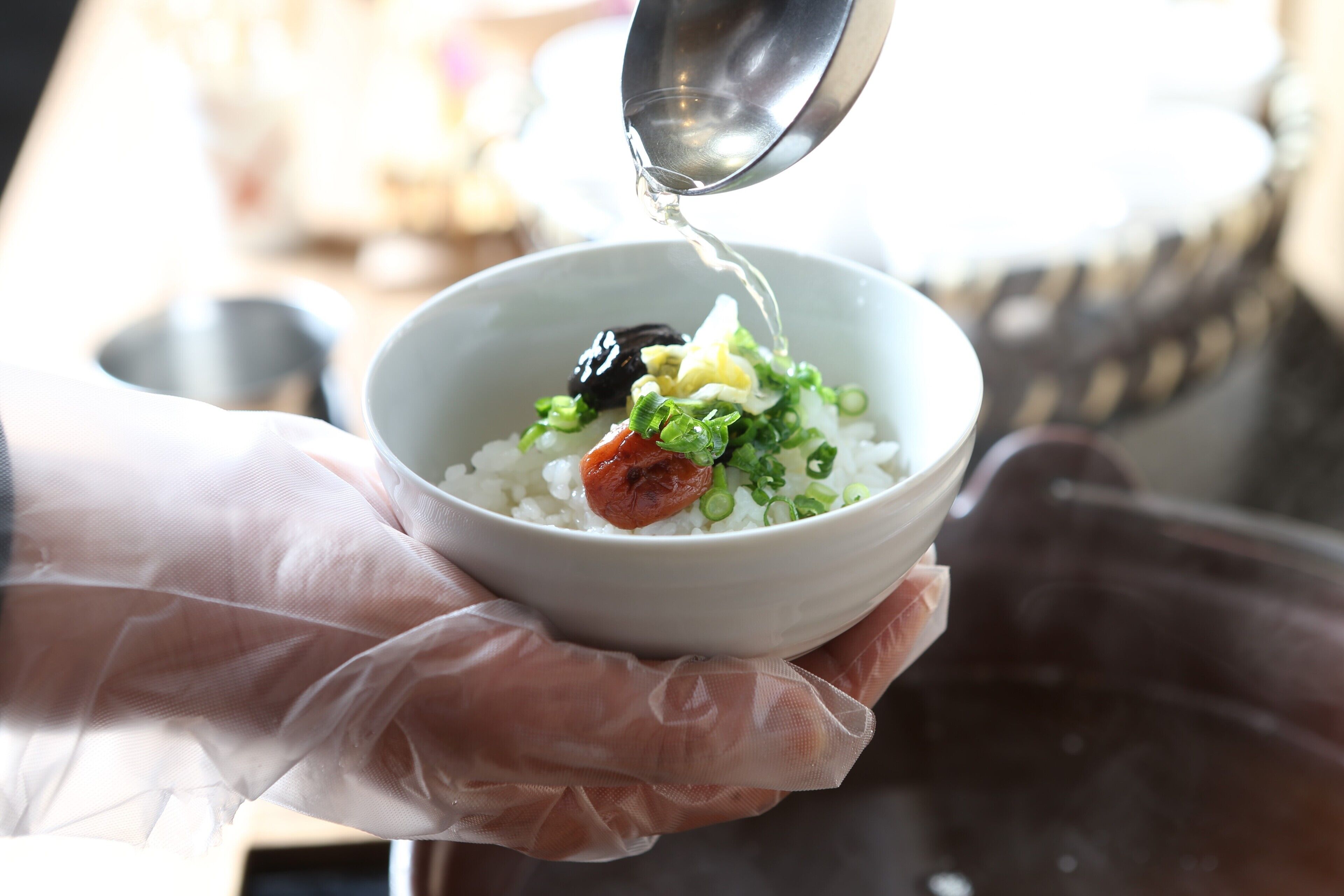 A person holding a bowl with rice, green onions, and a piece of meat, pouring liquid from a metal container.