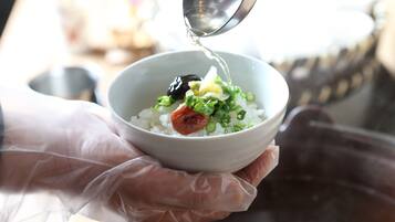 A person holding a bowl with rice, green onions, and a piece of meat, pouring liquid from a metal container.