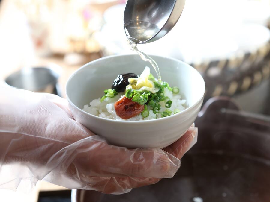 A person holding a bowl with rice, green onions, and a piece of meat, pouring liquid from a metal container.