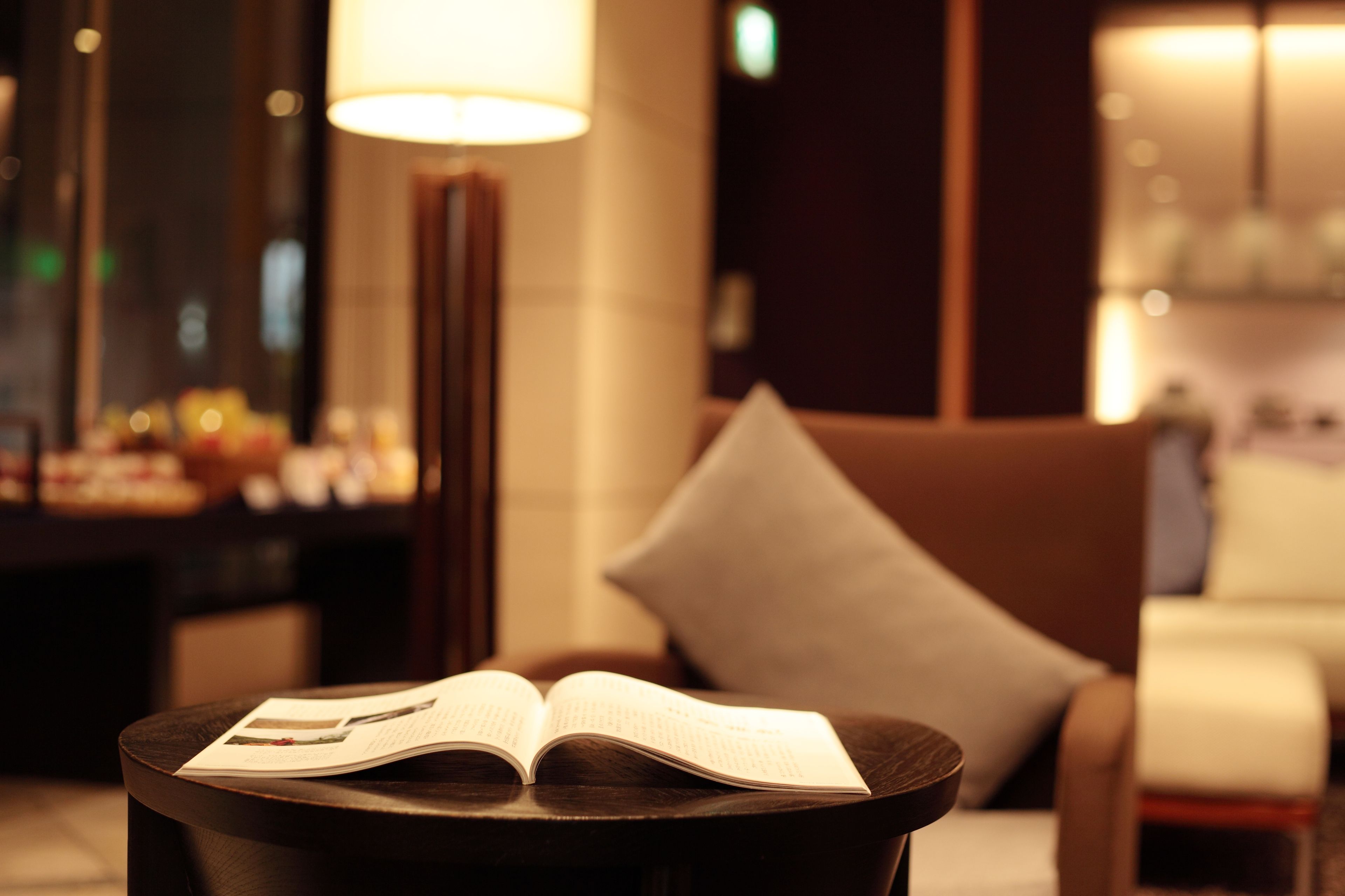 An open book on a small round table in a dimly lit room with a sofa and a lamp in the background.
