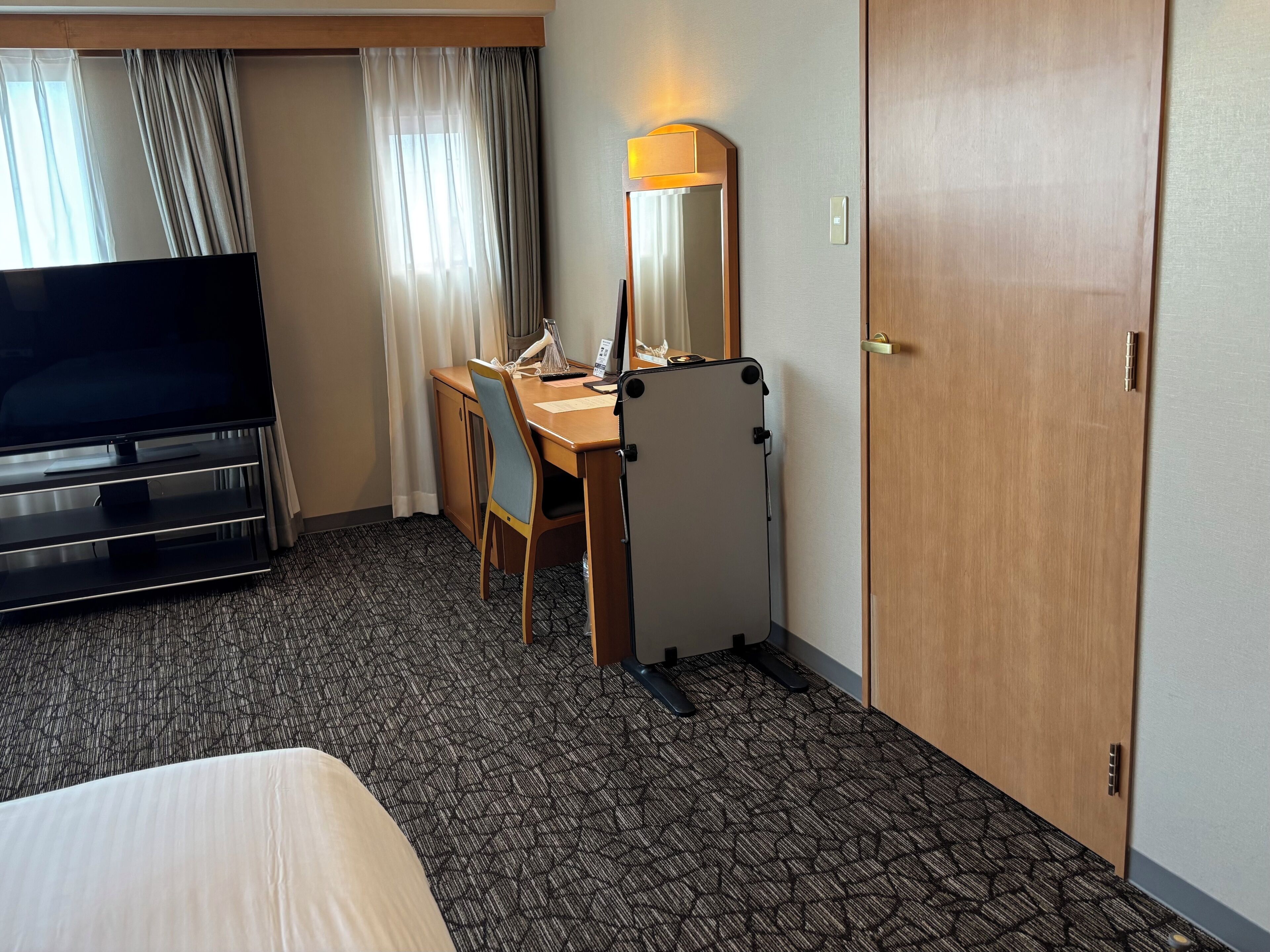A hotel room with a bed, a desk with a chair, a television, a mirror, and a door.