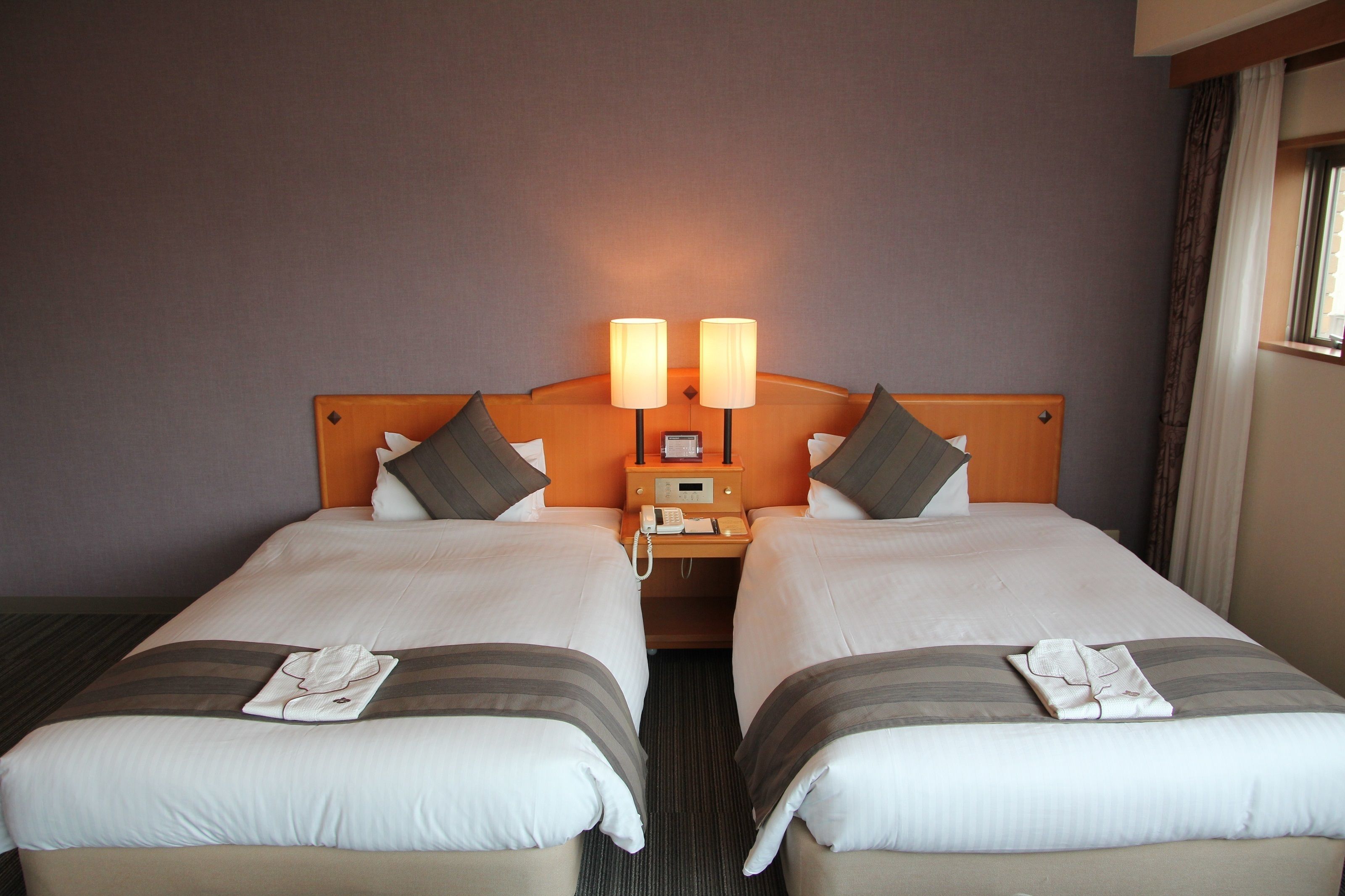 Habitación de hotel con dos camas, una mesita de noche con lámpara y una ventana con cortinas.