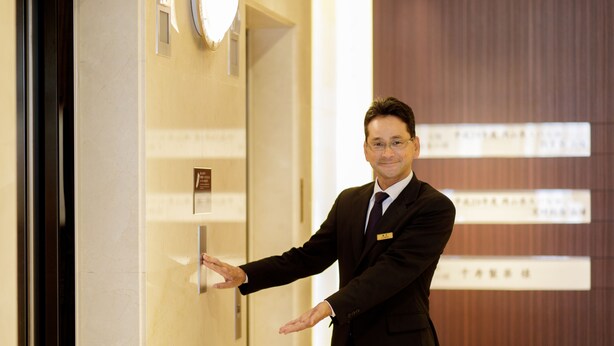 A man in a suit standing in a hallway with a door, a sign, and a wall with text. - Okayama Koraku Hotel (Okayama)
