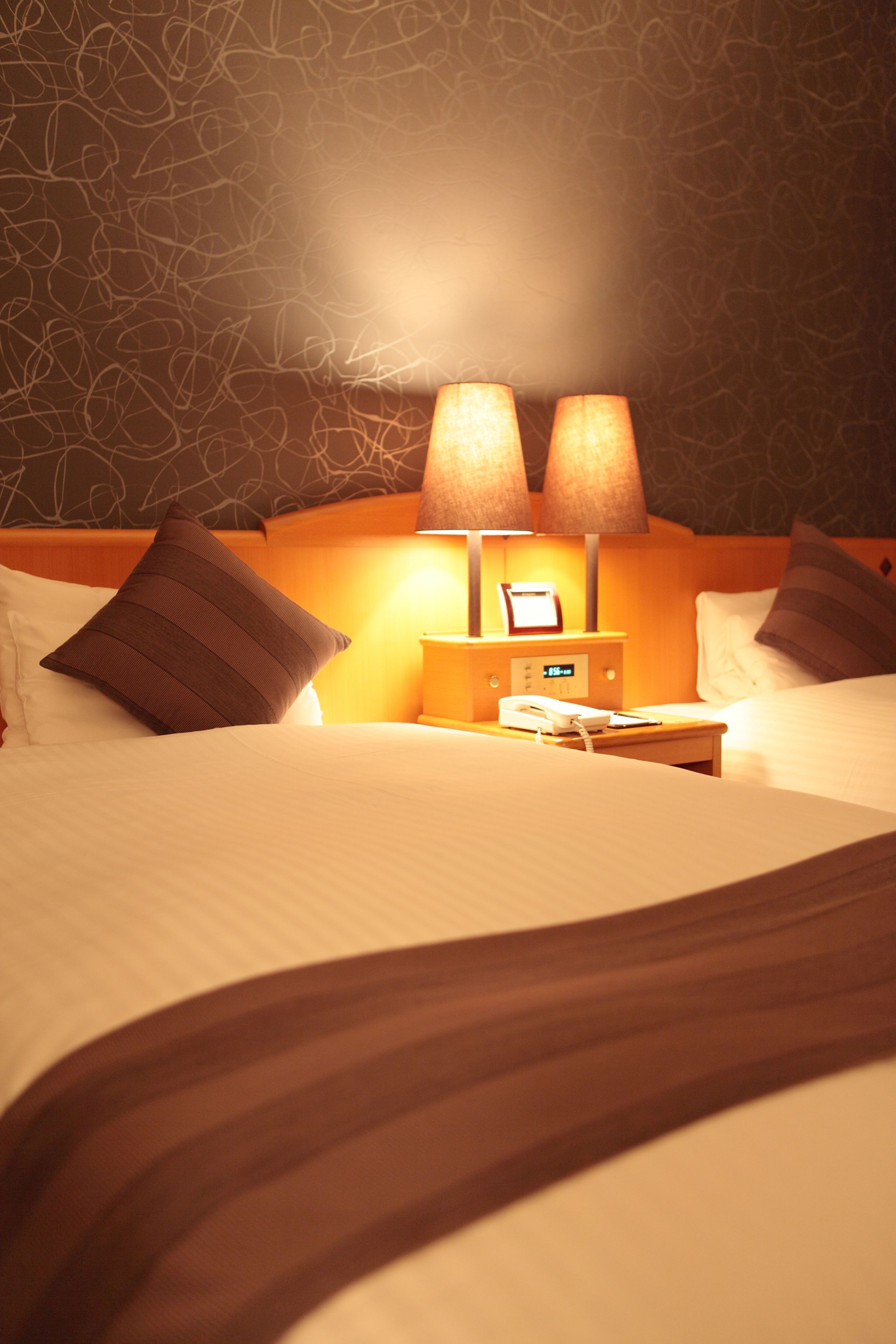 A hotel room with a neatly made bed, two bedside lamps, a nightstand with a phone, and a wall with a textured pattern.