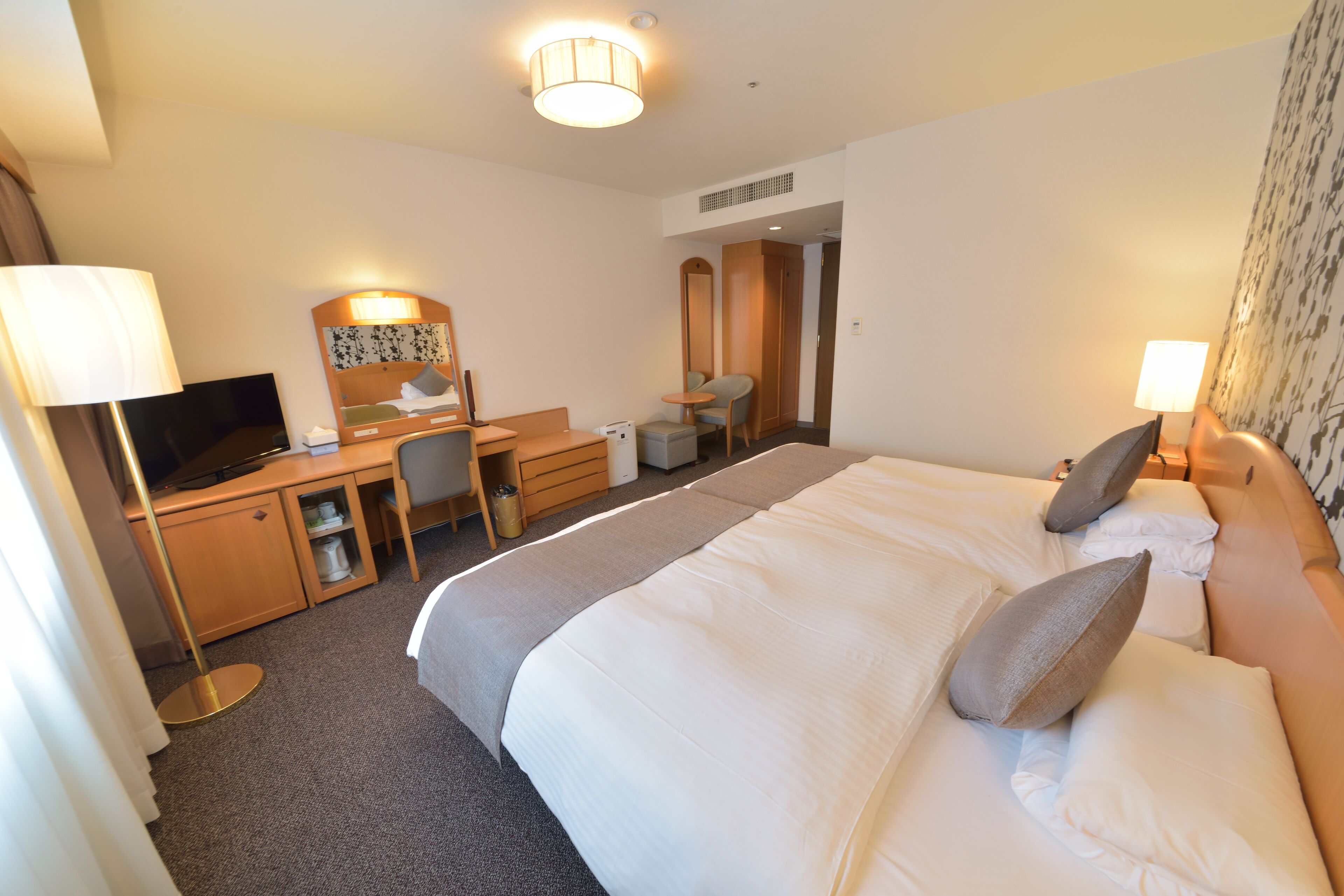 A hotel room with a large bed, a desk with a computer, a chair, a mirror, and a lamp.