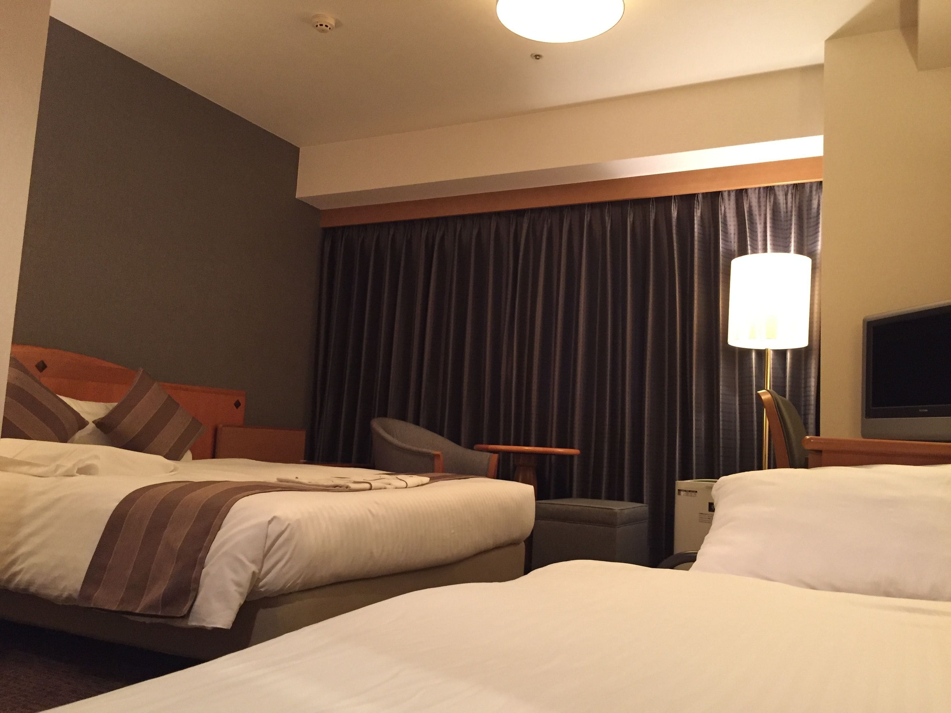 A hotel room with two beds, a desk, a chair, a TV, and a lamp.