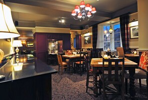 Un restaurante con poca luz, con mesas y sillas de madera, una zona de bar y una alfombra estampada.