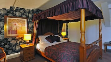 A four-poster bed with a dark canopy, a wooden nightstand with a lamp, a framed picture on the wall, and a floral wallpaper pattern.