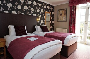 Habitación de hotel con dos camas, cabeceros oscuros, ropa de cama blanca, una manta roja, una mesita de noche, un espejo y una ventana con cortinas rojas.