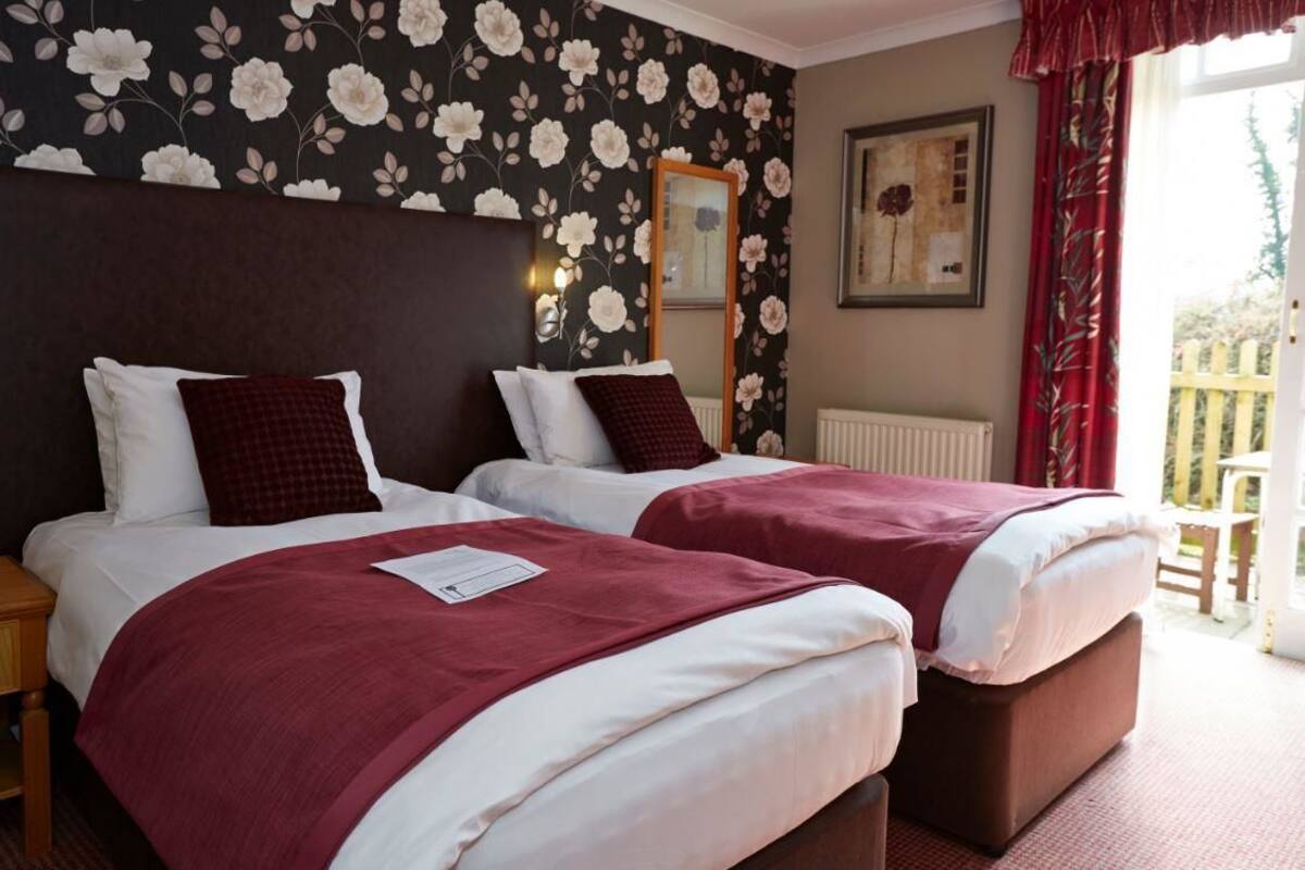 Habitación doble con dos camas, cabecera oscura, papel tapiz con motivos florales, un espejo y una ventana con cortinas rojas.