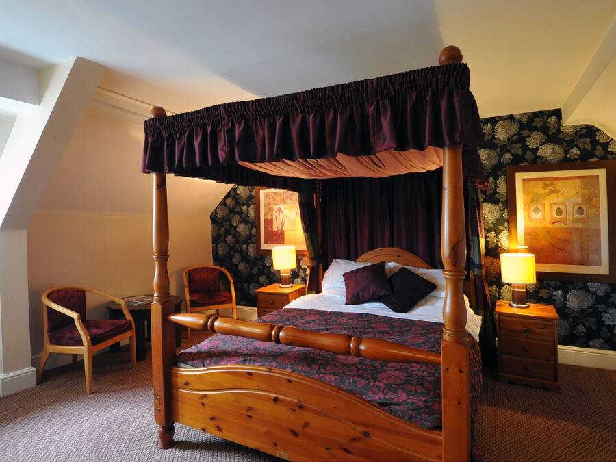 A four-poster bed with a canopy, a wooden nightstand with a lamp, a chair, and a framed picture on the wall.