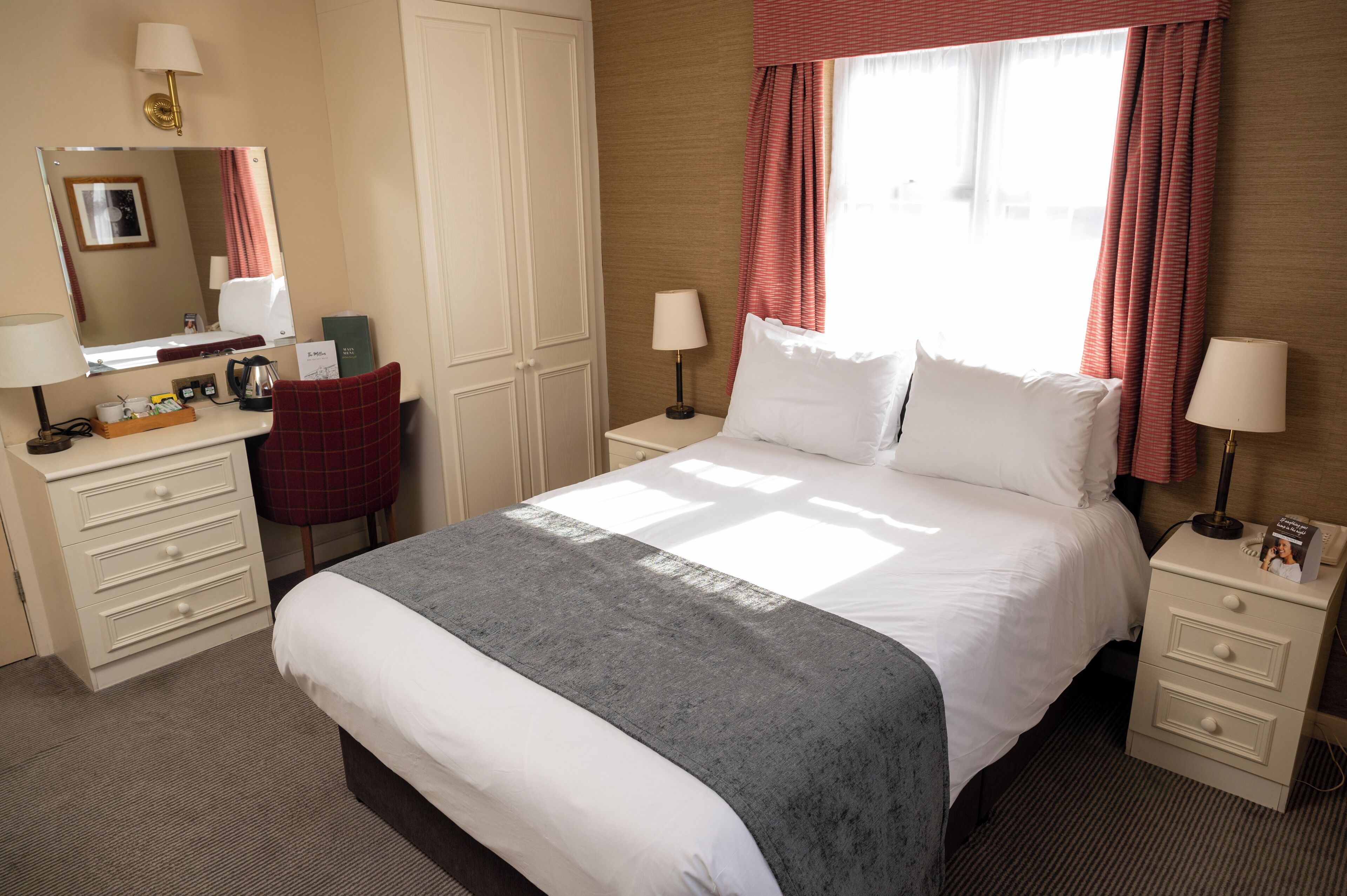 Ein Hotelzimmer mit Bett, Nachttischen, einem Schreibtisch mit Stuhl, einem Spiegel und einem fenster mit Vorhängen.