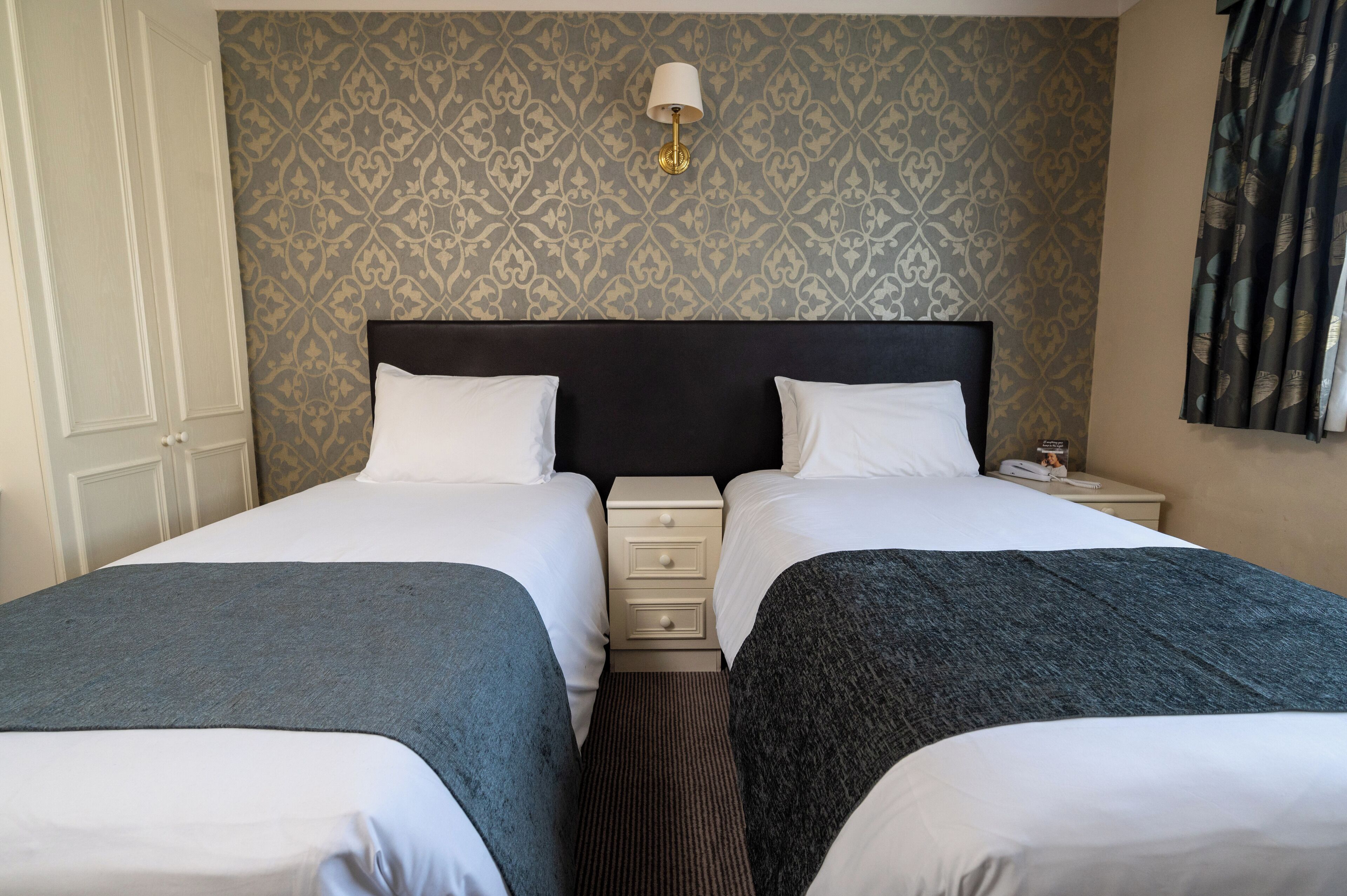 Habitación de hotel con dos camas, una mesita de noche, una lámpara y papel tapiz estampado.
