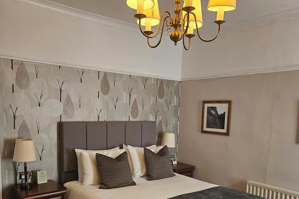 A hotel room with a large bed, two bedside tables, a chandelier, a patterned wallpaper, and a framed picture on the wall.