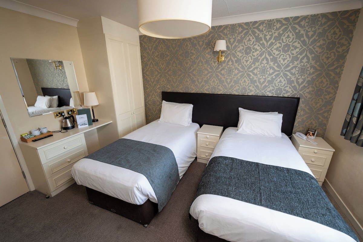 A hotel room with two beds, a dresser, a mirror, a lamp, and a patterned wallpaper.