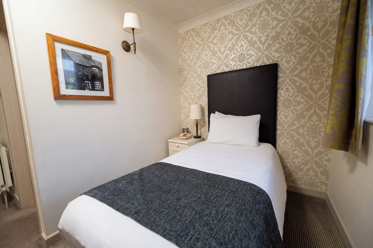 A hotel room with a large bed, a bedside table with a lamp, a framed picture on the wall, and patterned wallpaper.