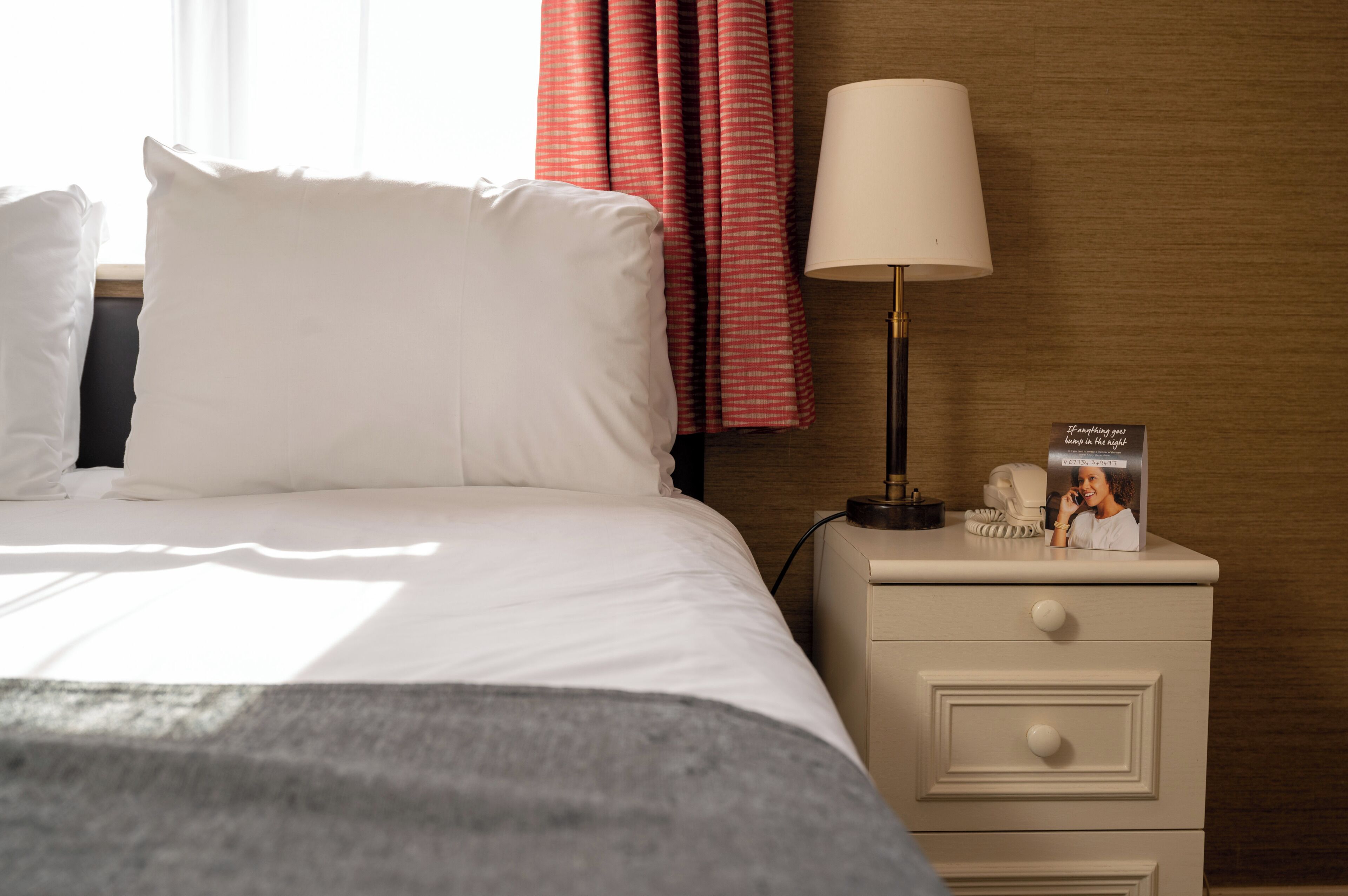 A neatly made bed with white linens, a bedside lamp, a nightstand with a phone and a magazine, and a window with curtains.