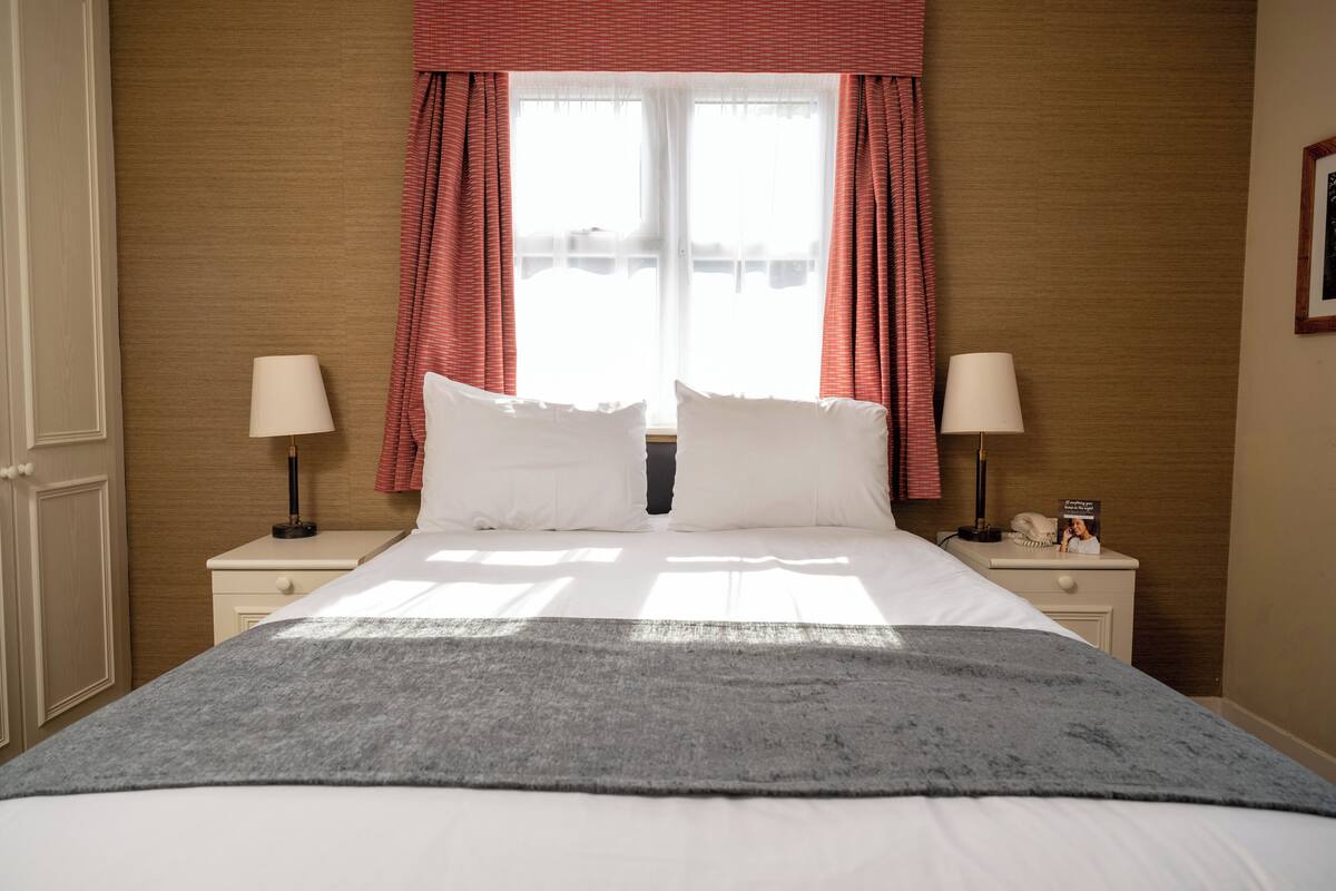 A neatly made bed with white linens, two bedside tables with lamps, and a window with red curtains.