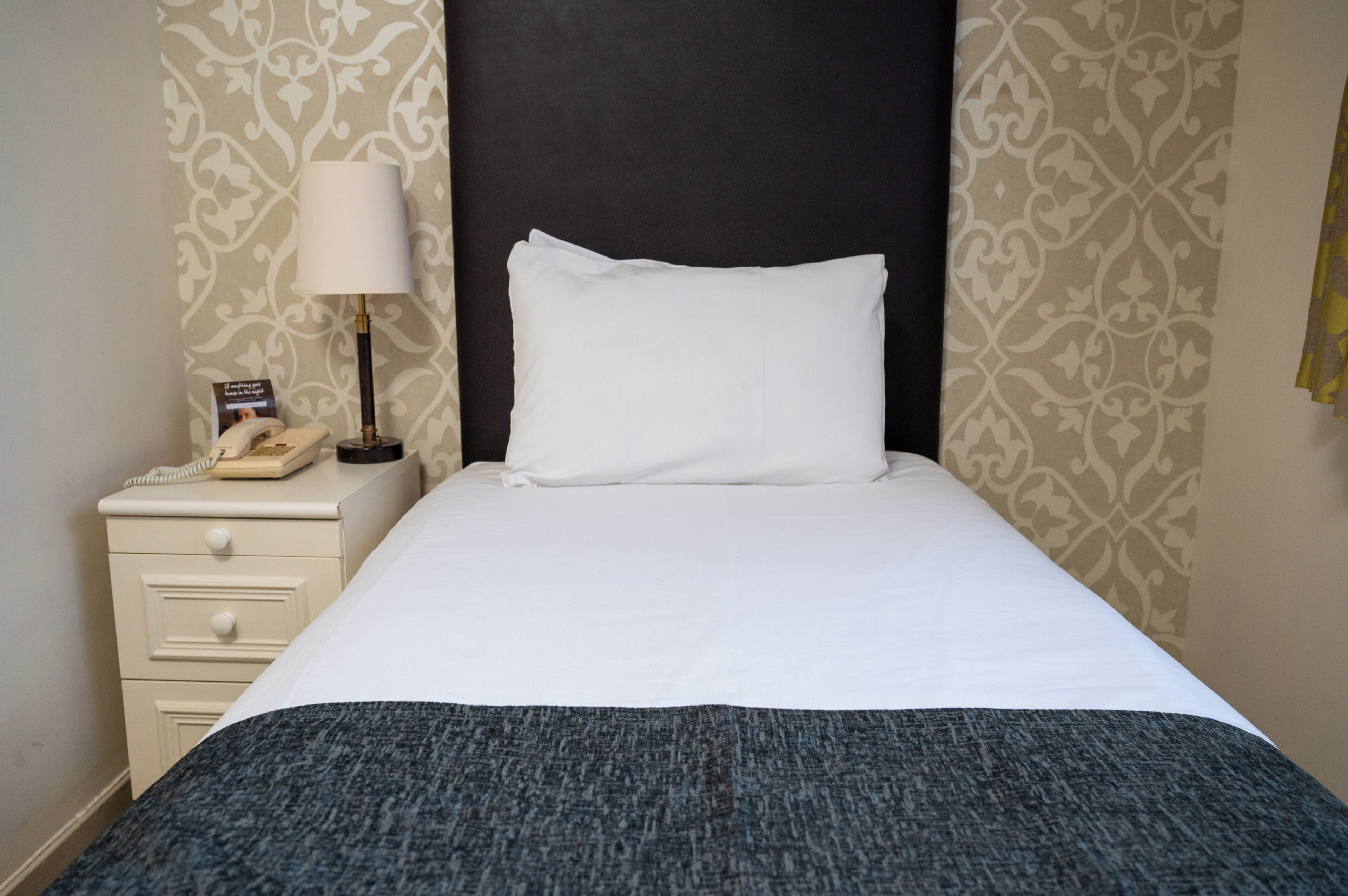 Ett hotellrum med en snyggt bäddad säng, en sänglampa, en telefon och en tapet med mönster.