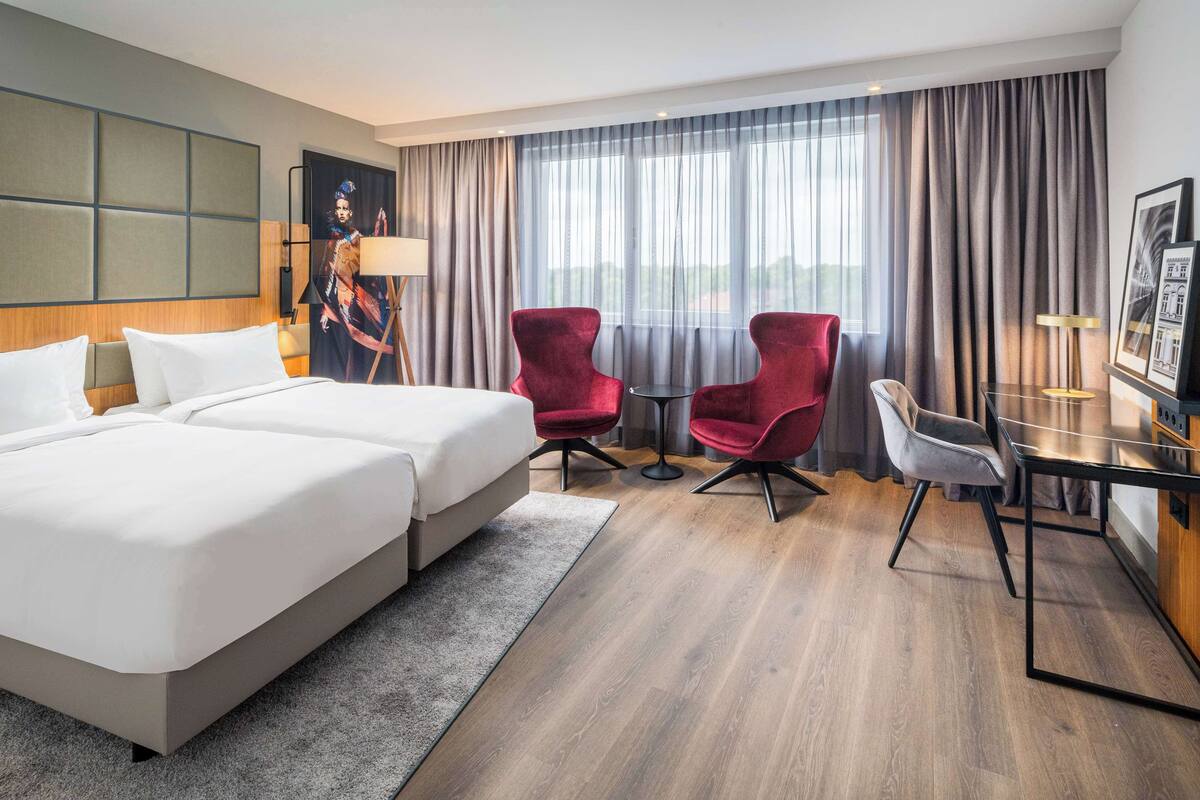 A hotel room with a large bed, two red armchairs, a small table, and a desk with a chair.
