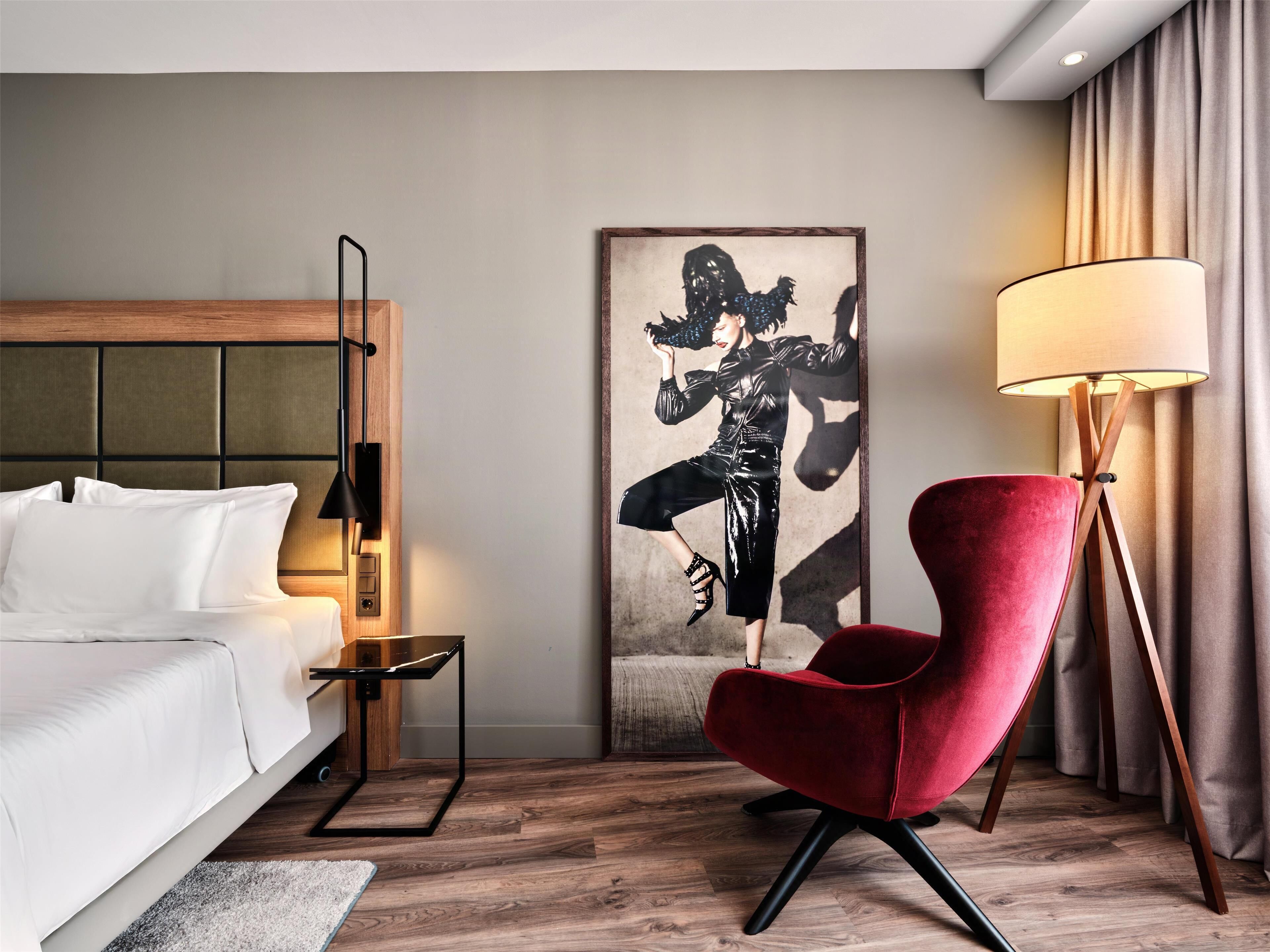 Ein modernes Hotelzimmer mit einem Bett, einem roten Sessel, einer Stehlampe und einem großen Kunstwerk an der Wand.