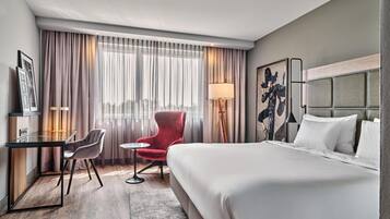 A hotel room with a bed, a desk, a chair, a lamp, and a large window with curtains.